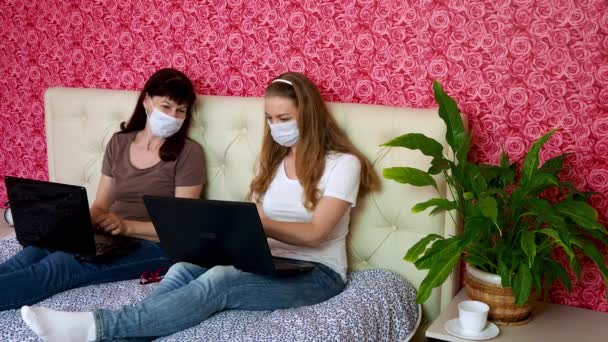 Távmunka otthonról. Két kaukázusi nő, egy fiatal szőke és egy felnőtt orvosi maszkban egy laptopon dolgoznak az ágyon, beszélgetnek és beszélgetnek a munkáról. Elektronikus oktatás. — Stock videók