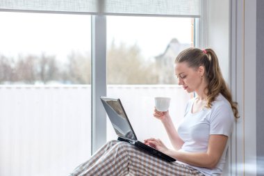Ev karantinası. Uzaktan kumandalı iş. Koronavirüsten koruma. Penceresinde bilgisayar ve bir fincan kahve olan beyaz bir kadın. Arkadaşlarla ve akrabalarla çevrimiçi iletişim kurmak ve öğrenmek.