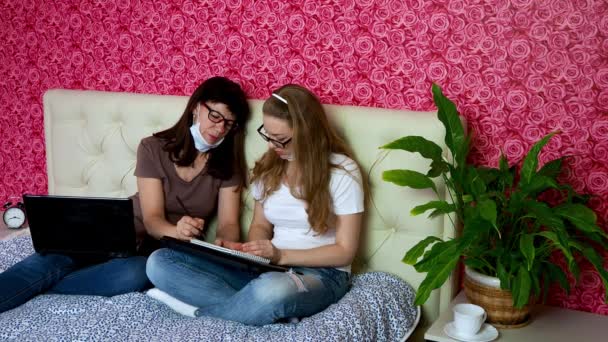 Op afstand werken. Een studente wordt thuis onderwezen met een leraar. Twee blanke vrouwen, moeder en dochter met beschermende maskers, schrijven in een notitieboekje, maken huiswerk, hebben plezier, lachen. Zelfisolatie in quarantaine. — Stockvideo