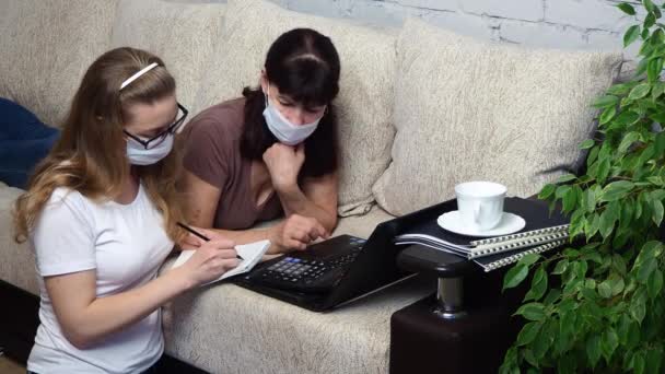 Travail à distance depuis la maison. Deux femmes portant des masques médicaux protecteurs travaillant ensemble sur un ordinateur portable sur le canapé du salon, prenant des notes dans un carnet. E-éducation. Volgodonsk, Russie-07 avril 2020 . — Video