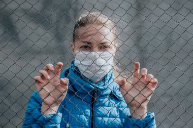 Koruyucu tıbbi maskeli beyaz bir kız metal bir çitin arkasında duruyor ve üzgün görünüyor. Ayrılmak istiyor. Karantina sırasında katı ev izolasyonu. Sokağa çıkma yasağı var. Kendini koruma..