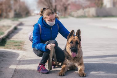 Koruyucu tıbbi maskeli bir kız sokakta bir köpeği gezdiriyor. Karantina sırasında bir evcil hayvanla vakit geçirmek. Temiz havada bir Alman çobanla yürüyün. Kişisel izolasyon ve koruma modu.