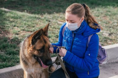 Koruyucu tıbbi maskeli bir kız sokakta bir köpeği gezdiriyor. Karantina sırasında bir evcil hayvanla vakit geçirmek. Temiz havada bir Alman çobanla yürüyün. Kişisel izolasyon ve koruma modu.