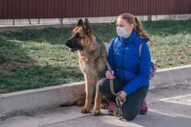 Koruyucu tıbbi maskeli bir kız sokakta bir köpeği gezdiriyor. Karantina sırasında bir evcil hayvanla vakit geçirmek. Temiz havada bir Alman çobanla yürüyün. Kişisel izolasyon ve koruma modu.