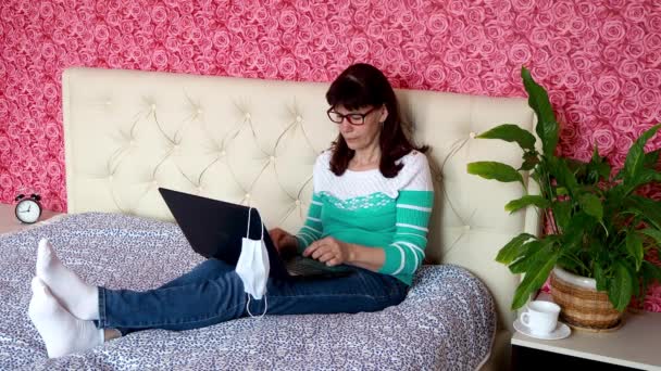 Werk op afstand van huis. Een volwassen blanke vrouw werkt op een laptop in haar slaapkamer op een bed met een beschermend medisch masker. Het concept van het bedrijfsleven en e-onderwijs. Zelfisolatie in quarantaine. — Stockvideo