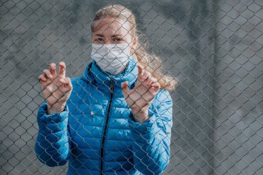 Koruyucu tıbbi maskeli beyaz bir kız metal bir çitin arkasında duruyor ve üzgün görünüyor. Ayrılmak istiyor. Karantina sırasında katı ev izolasyonu. Sokağa çıkma yasağı var. Kendini koruma..