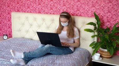 Uzaktan kumandalı iş. Beyaz güzel bir kız yatak odasında dizüstü bilgisayarla koruyucu bir maske takıp kahve içiyor. İş ve e-eğitim. Karantinada kendini izole etme.
