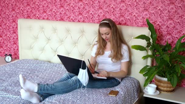 Távmunka otthon. Egy gyönyörű kaukázusi lány jegyzeteket ír egy jegyzetfüzetbe a hálószobájában az ágyon egy védő orvosi maszkban. Az üzleti élet és az e-oktatás fogalma. Önképzés a karanténban. — Stock videók