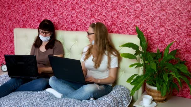 Evden uzak bir iş. Koruyucu tıbbi maske takmış iki beyaz kadın yatak odasındaki dizüstü bilgisayarda çalışıyor, neşeyle konuşuyor, gülümsüyor ve işten bahsediyorlar. E-eğitim. — Stok video