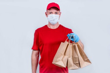 Güvenli teslimat kavramı. Kırmızı üniformalı, koruyucu tıbbi maskeli ve eldivenli bir kurye, koronavirüs salgını sırasında karantinaya alınması gereken siparişleri ileten bir kese kağıdı tutuyor.