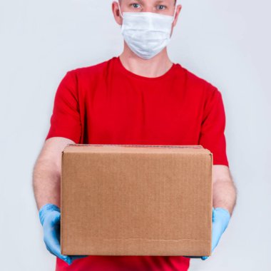 Güvenli teslimat kavramı. Kırmızı üniformalı, koruyucu tıbbi maskeli ve eldivenli bir kurye karton bir kutu tutuyor. Coronavirus salgını sırasında karantinaya alınması yasak.