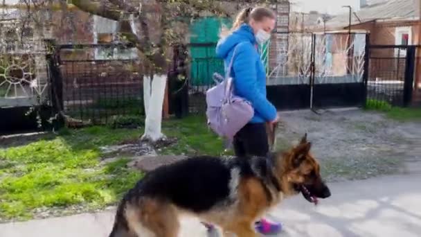 Ein Mädchen mit einer medizinischen Schutzmaske geht mit einem Hund auf der Straße spazieren. Freizeit mit einem Haustier während der Quarantäne. Wandern Sie mit einem Schäferhund an der frischen Luft. Selbstisolation und Schutzmodus. — Stockvideo