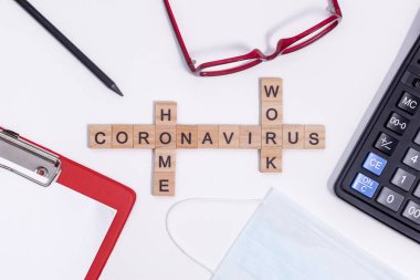 Tahta harflerden gelen mesaj Coronavirus evden çalışıyor. Masaüstünde ofis malzemeleri, hesap makinesi, tablet, gözlük, kalem ve koruyucu tıbbi maske var. Beyaz arkaplanda düz bir düzen