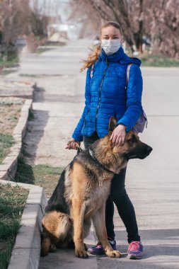 Koruyucu tıbbi maskeli bir kız sokakta bir köpeği gezdiriyor. Karantina sırasında bir evcil hayvanla vakit geçirmek. Temiz havada bir Alman çobanla yürüyün. Kişisel izolasyon ve koruma modu.