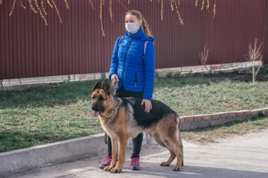 Koruyucu tıbbi maskeli bir kız sokakta bir köpeği gezdiriyor. Karantina sırasında bir evcil hayvanla vakit geçirmek. Temiz havada bir Alman çobanla yürüyün. Kişisel izolasyon ve koruma modu.