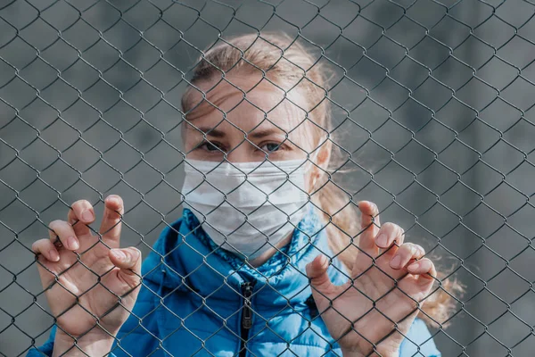 Koruyucu tıbbi maskeli beyaz bir kız metal bir çitin arkasında duruyor ve üzgün görünüyor. Ayrılmak istiyor. Karantina sırasında katı ev izolasyonu. Sokağa çıkma yasağı var. Kendini koruma.