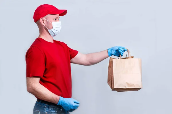 安全な配達の概念 赤い制服と保護医療マスクと手袋の宅配便は コロナウイルスパンデミックの間に隔離された紙袋 非接触配達を保持しています — ストック写真