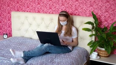 Uzaktan kumandalı iş. Beyaz güzel bir kız yatak odasında dizüstü bilgisayarla koruyucu bir maske takıp kahve içiyor. İş ve e-eğitim. Karantinada kendini izole etme.