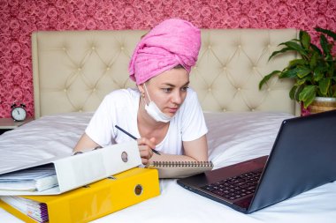 Ev karantinasından uzak bir çalışma. Maskeli, ödevlerini yapan beyaz bir kız. Dizüstü bilgisayarına bakar ve bir not defterine not alır. E-eğitim. Salgın sırasında çevrimiçi eğitim