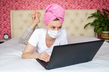 Ev karantinasından uzak bir çalışma. Maskeli bir yatağın üstünde dizüstü bilgisayar üzerinde çalışan beyaz bir kız blogcu. Uzaktan iletişim, online alışveriş, salgın sırasında ev siparişleri