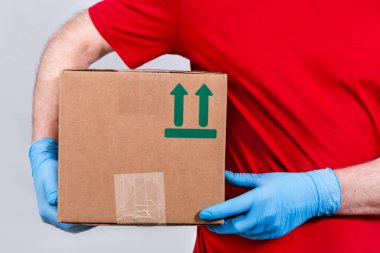 Güvenli teslimat kavramı yakın planda. Kırmızı üniformalı ve koruyucu tıbbi eldivenli bir kurye elinde karton bir kutu tutuyor. Coronavirus salgını sırasında karantinaya alınması gereken siparişlerin hiç bir şekilde iletilmiyor.