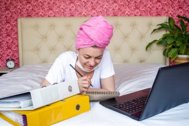 Ev karantinasından uzak bir çalışma. Maskeli, ödevlerini yapan beyaz bir kız. Dizüstü bilgisayarına bakar ve bir not defterine not alır. E-eğitim. Salgın sırasında çevrimiçi eğitim