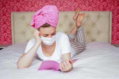 Mesafe öğrenme. Ev karantinası. Koruyucu tıbbi maske takmış, kafasında havlu olan beyaz bir kız yatakta kitap okuyor. Salgın sırasında eğlence ve hobiler. Kendi kendini eğitme