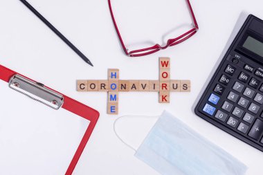 Tahta harflerden gelen mesaj Coronavirus evden çalışıyor. Masaüstünde ofis malzemeleri, hesap makinesi, tablet, gözlük, kalem ve koruyucu tıbbi maske var. Beyaz arkaplanda düz bir düzen