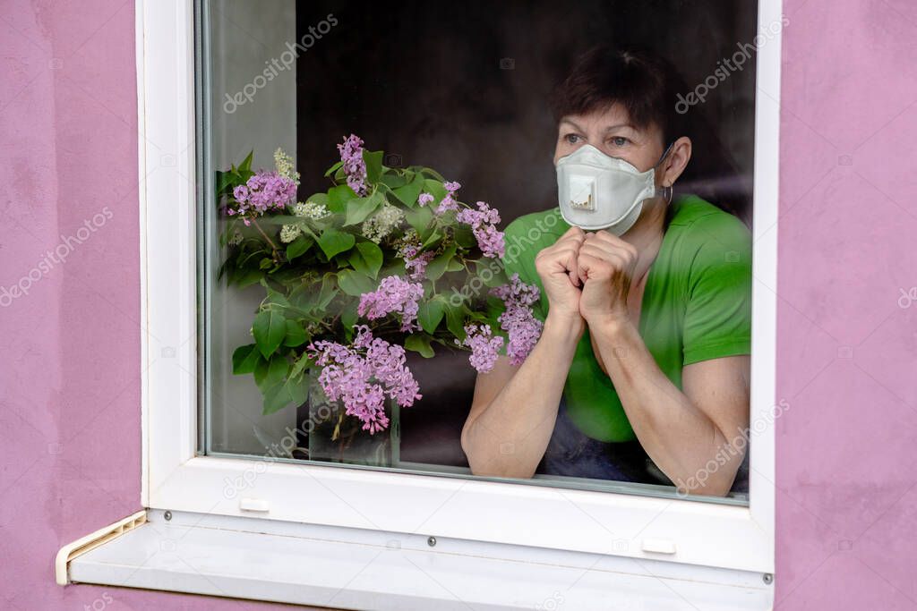 A sad older woman in a protective mask looks out the window, waiting for the end of self-isolation. The concept of coronavirus quarantine stay at home and social distance. the elderly lifestyle