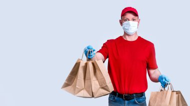 Pankart. Güvenli teslimat. Kırmızı üniformalı, koruyucu maskeli ve eldivenli bir kurye büyük bir sipariş, bir sürü kese kağıdı ve karantinaya konulamaz yiyecek taşır. Gönüllü bağışı. Boşluğu kopyala