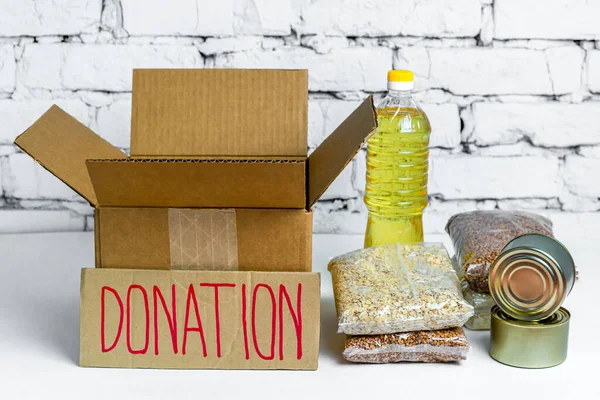 Food in cardboard donation box on a white background. Anti-crisis stock of essential goods for period of quarantine isolation. Food delivery, coronavirus. The shortage of food. Sign with text.