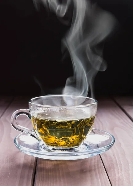 A transparent cup of hot green tea and steam on a dark background.