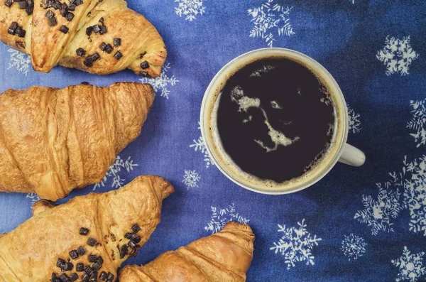 Interesting idea for serving coffee with a sweet snack. Coffee with croissants on a blue winter background.