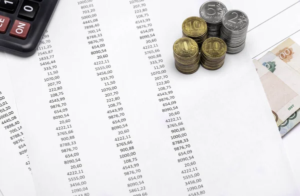 Close-up printed numbers on white paper, on which lies the calculator, coins of different values of rubles and next to paper bills.