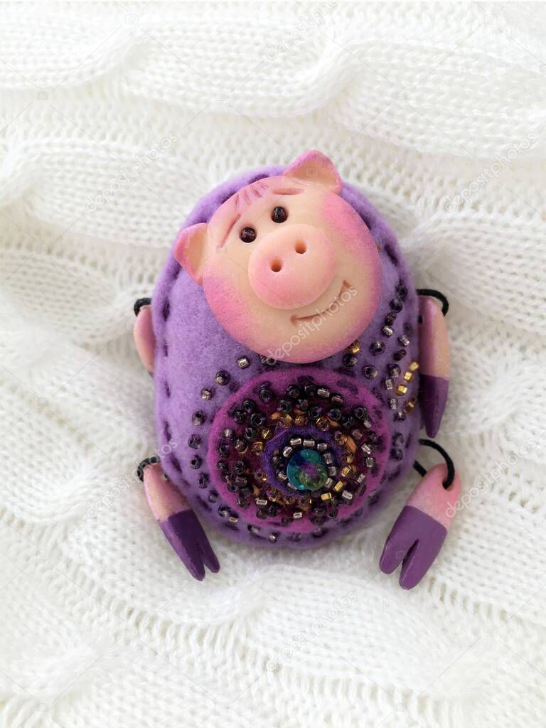 Toy pig made of polymer clay and purple felt, decorated with beads on a white plaid background