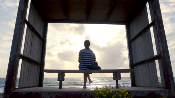 Κορίτσι σε ένα παγκάκι στην παραλία το ηλιοβασίλεμα — Αρχείο Βίντεο