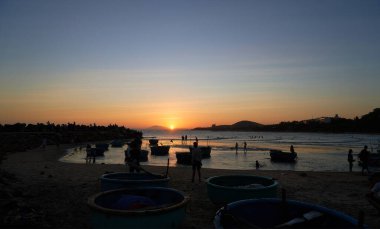 İnsanlar gün batımında kıyıda yuvarlak teknelerle dinleniyor.         