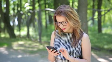 Parkta akıllı telefon kullanan gözlüklü mutlu genç bir kadın.