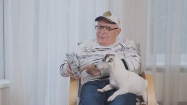 Gözlüklü ve şapkalı emekli koltukta tablet köpeğe gösteriyor