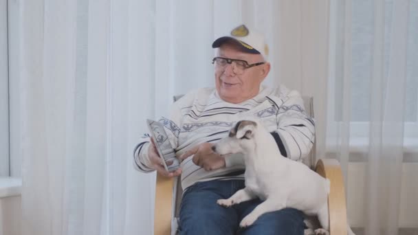 Pensionné dans les lunettes et le chapeau montre tablette au chien dans le fauteuil — Video