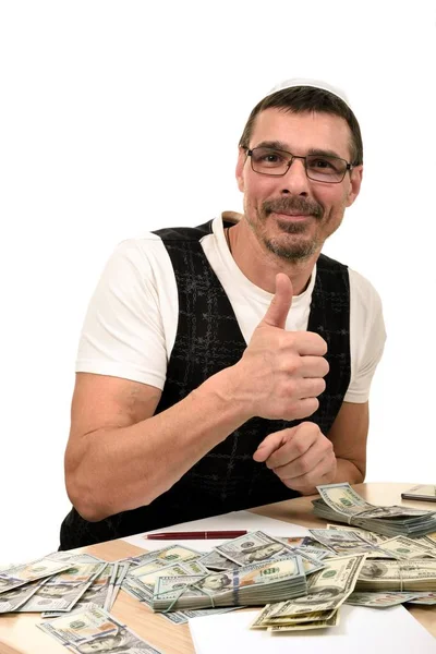 一个快乐的人在一个犹太帽和背心的肖像坐在桌子上 有很多钱 并显示拇指 — 图库照片