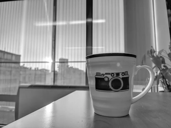 Black and white retro style old camera image print on a white mug placed on a desk while working on the job and drinking coffee — Stock Photo, Image