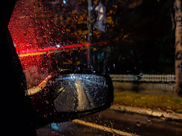 Amazing atmosphere during night while driving on a rainy street with closeup of red car street lights and drops of rain in focus on a car window glass with side rear view mirror — Stock Photo, Image