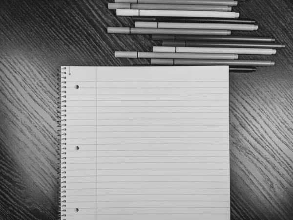 Top view image of empty notebook paper sheet with lines, holes and spiral on wooden desk background and colorful pens above in black and white — Stock Photo, Image