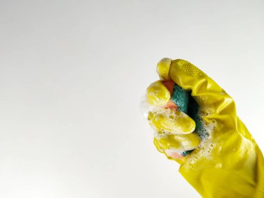 Sarı kauçuk eldivenin beyaz arka planında, köpük dolu bir sünger, deterjan ve temizlik için sabun tutarken çekilmiş yakın plan ve üst manzara.
