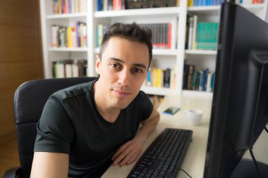 Odasında gülümseyen ve mutlu bir adam bilgisayarın başında oturmuş kameraya bakıyor, tişört giyiyor. Orta çekim.