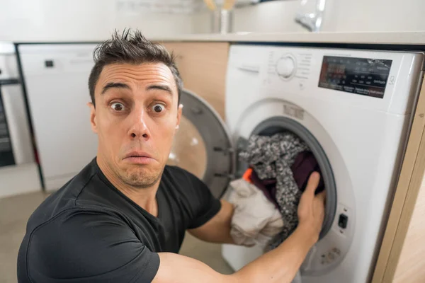 Man putting clothes in the washing machine, in the kitchen, worried because he has to wash a lot of clothes. Close up.