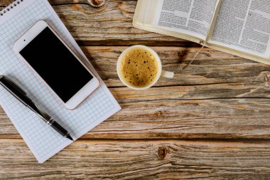 Los Angeles CA US 16 MAY 2020: Morning study with open Holy Bible with black coffee cup on smartphone and pen over spiral notepad on wooden background clipart