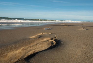 Kumlu sahilde temiz ayak izlerinin yakınlaşması. Arka planda dalgalar ve deniz var.