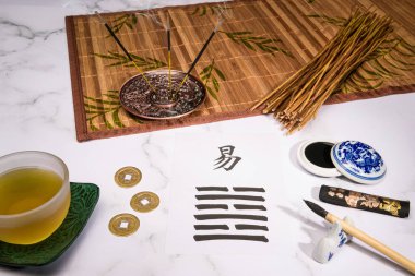 Çinli bir mürekkep fırçasıyla yazılmış 63. Hexagram (Tamamlandıktan sonra / Chi Chi) ile I Ching düzenlemesini kapat. Bir fincan yeşil çay, tütsü çubukları, kiraz sapları, I Ching paraları, bambu hasırı ve beyaz mermerin üzerine Çin mürekkebi çubuğu..